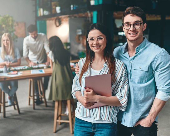 A smiling couple stands in a modern work environment. The woman is holding a notebook while the man embraces her. In the background, several people are engaged at a table.